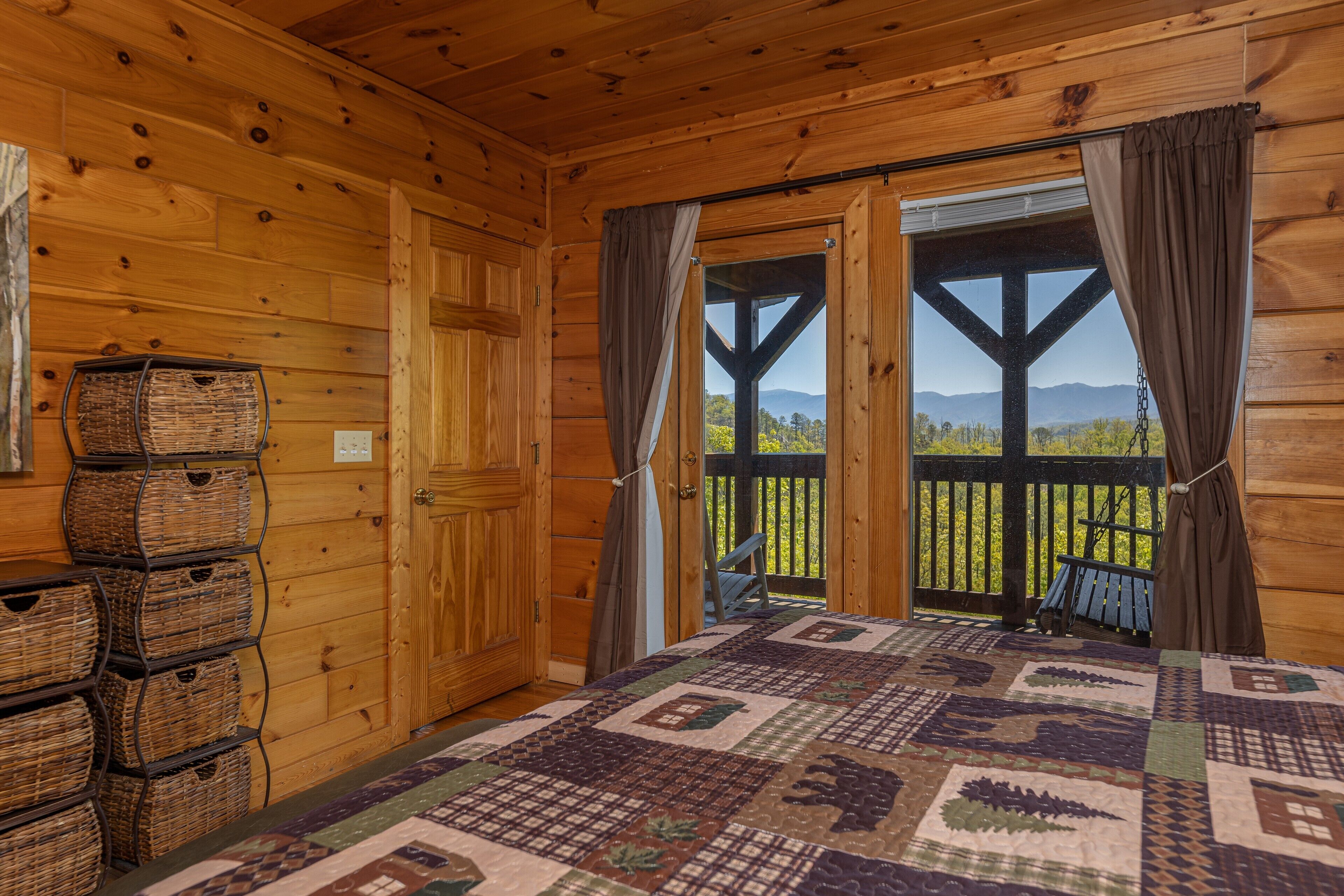 A wooden cabin room with a large bed, a view of mountains, and wicker baskets on a shelf.