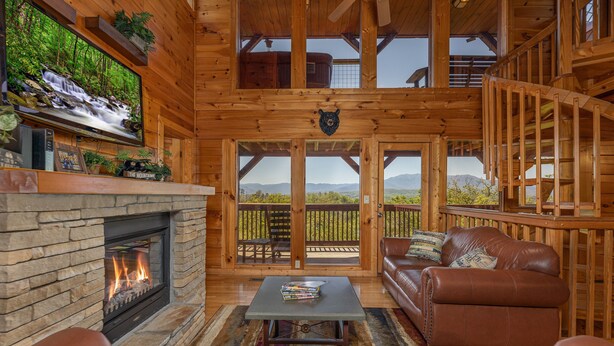 Interior acogedor de una cabaña con chimenea de piedra, muebles de cuero, un televisor montado en la pared y vistas a las montañas a través de grandes ventanales.