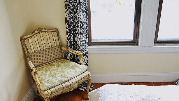 A room with a wooden floor, a white bed, a wicker chair with a floral cushion, and a window with a black and white patterned curtain. - The King's Room at TPH - Boston (Boston)