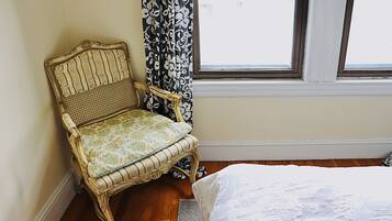 A room with a wooden floor, a white bed, a wicker chair with a floral cushion, and a window with a black and white patterned curtain.