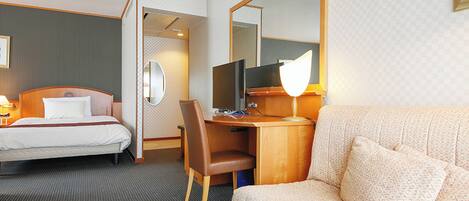 A hotel room with a bed, a desk with a computer, a chair, a sofa, a mirror, and a lamp.