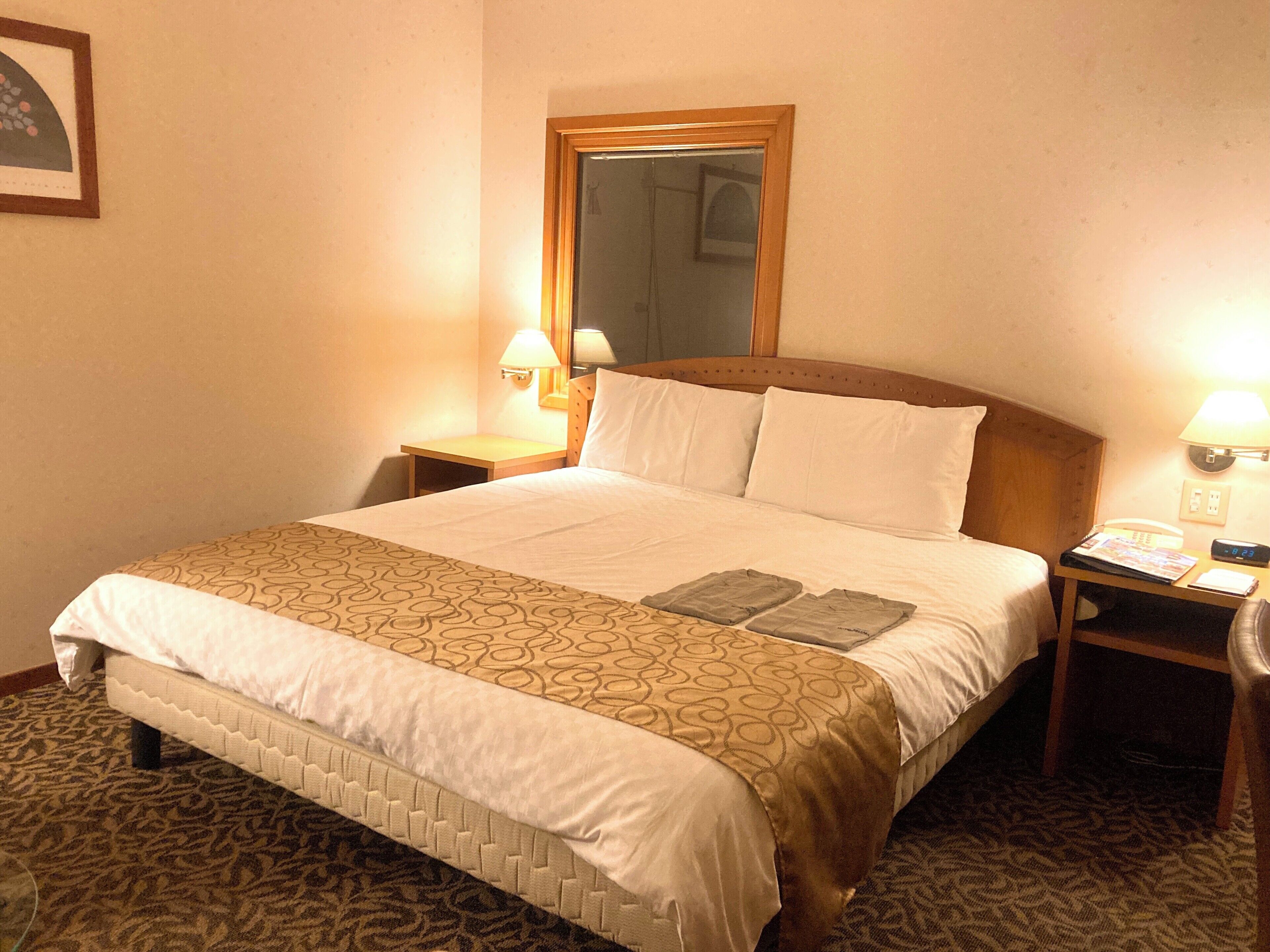 A hotel room with a large bed, two bedside tables, a nightstand, a mirror, and a wall-mounted lamp.