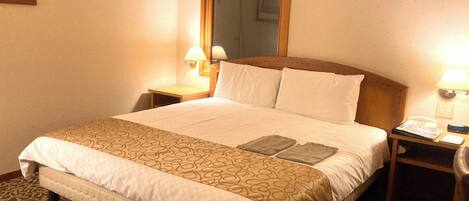 A hotel room with a large bed, two bedside tables, a nightstand, a mirror, and a wall-mounted lamp.