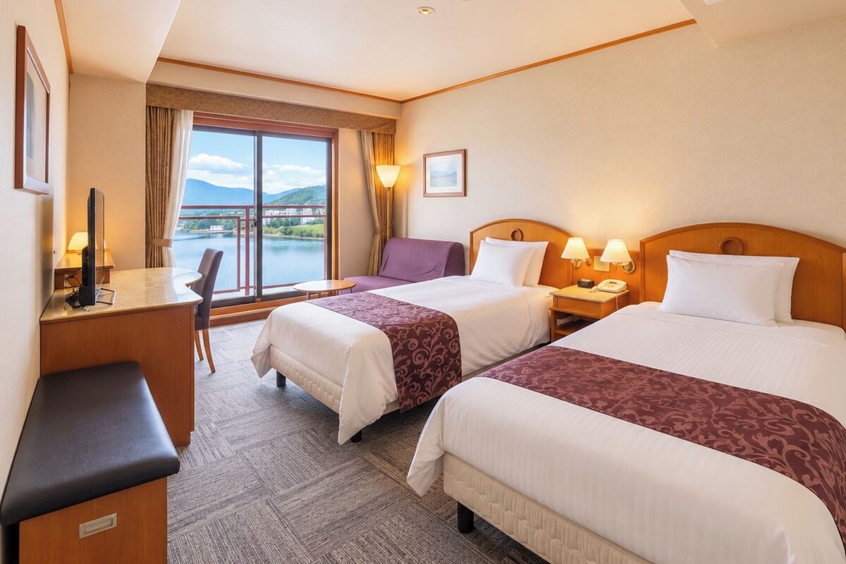 A hotel room with two beds, a desk, a chair, a lamp, and a view of the outdoors.