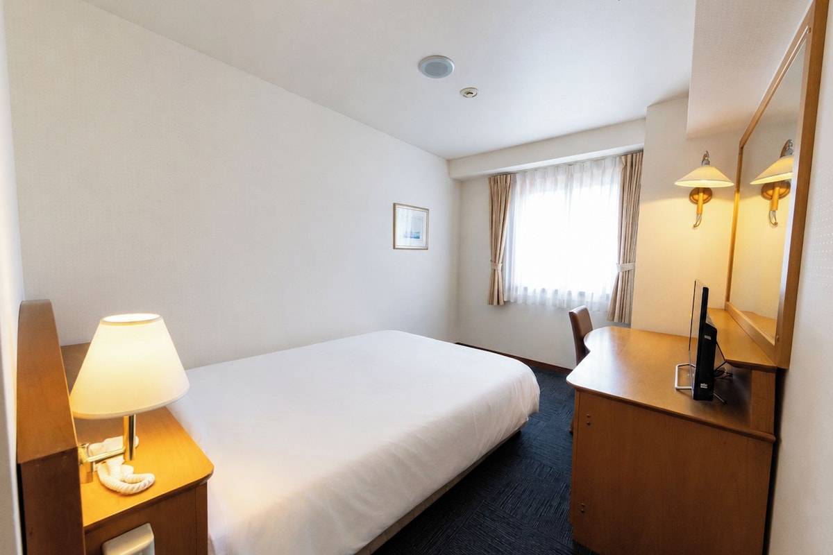 A hotel room with a single bed, a bedside table with a lamp, a desk with a computer, and a mirror.