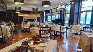 A restaurant with multiple dining tables set for guests, featuring a piano in the background and large windows allowing natural light.