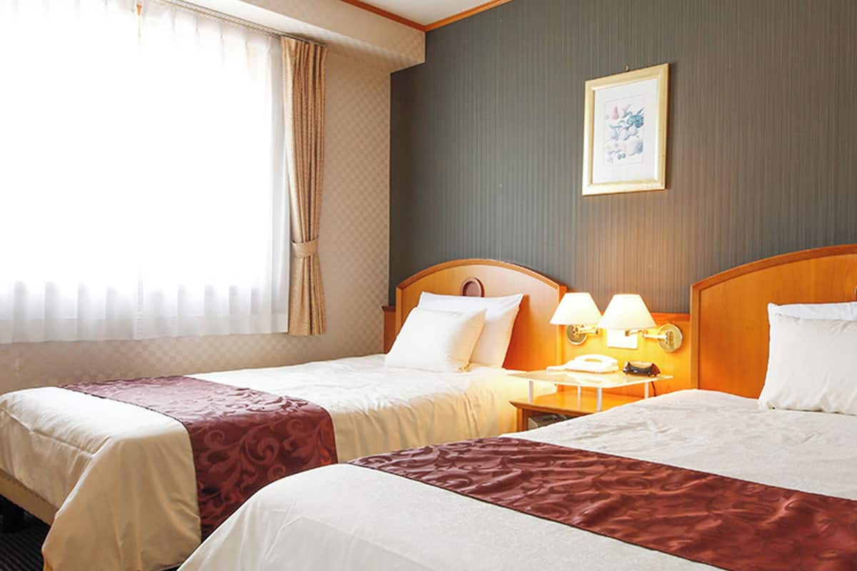 Hotellihuone, jossa on kaksi sänkyä, yöpöytä lamppuineen, seinällä oleva kehystetty taulu ja ikkuna, jossa on verhot.