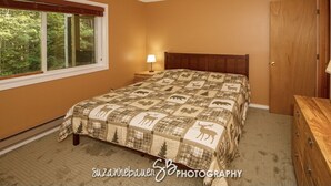 Quarto com cama, mesa de cabeceira, cômoda e janela com vista para árvores.