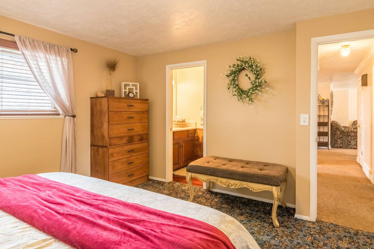 Une chambre à coucher comprenant un lit, une commode, un banc, une porte donnant sur une salle de bain et un couloir menant à une autre pièce.