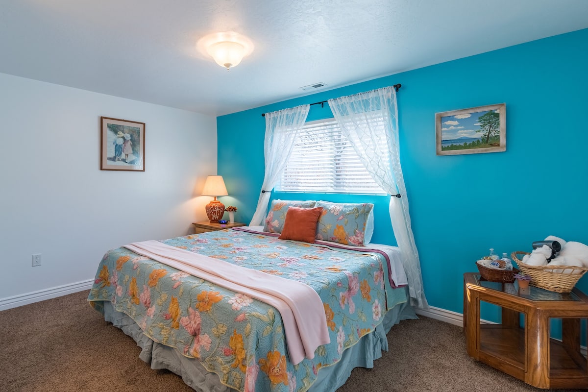 Une chambre à coucher avec une couvre-lit à motifs floraux, une lampe de chevet, une fenêtre avec des rideaux légers et une petite table d’appoint en bois.