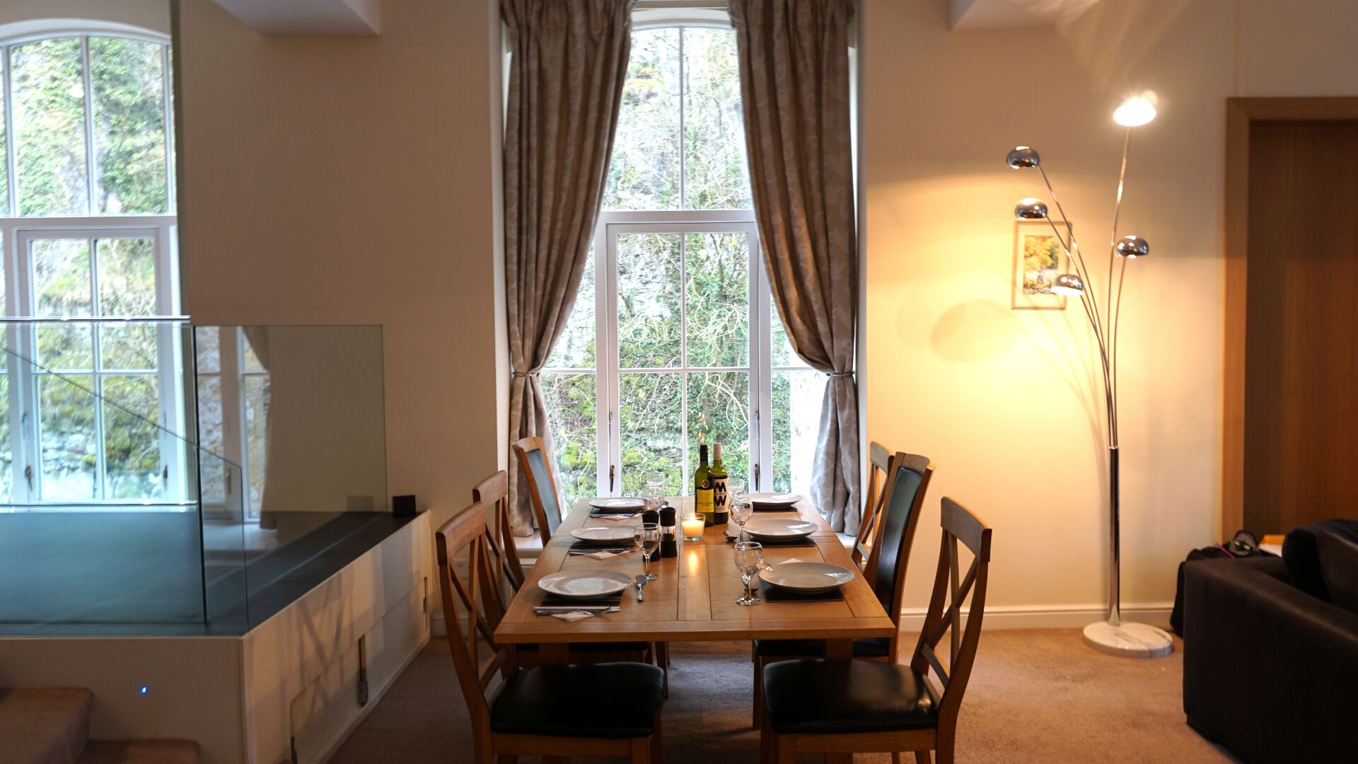 Comedor con mesa de madera dispuesta para cuatro personas, una vela y una botella de vino. Cortinas en las ventanas, una lámpara de pie y vistas al verdor exterior.