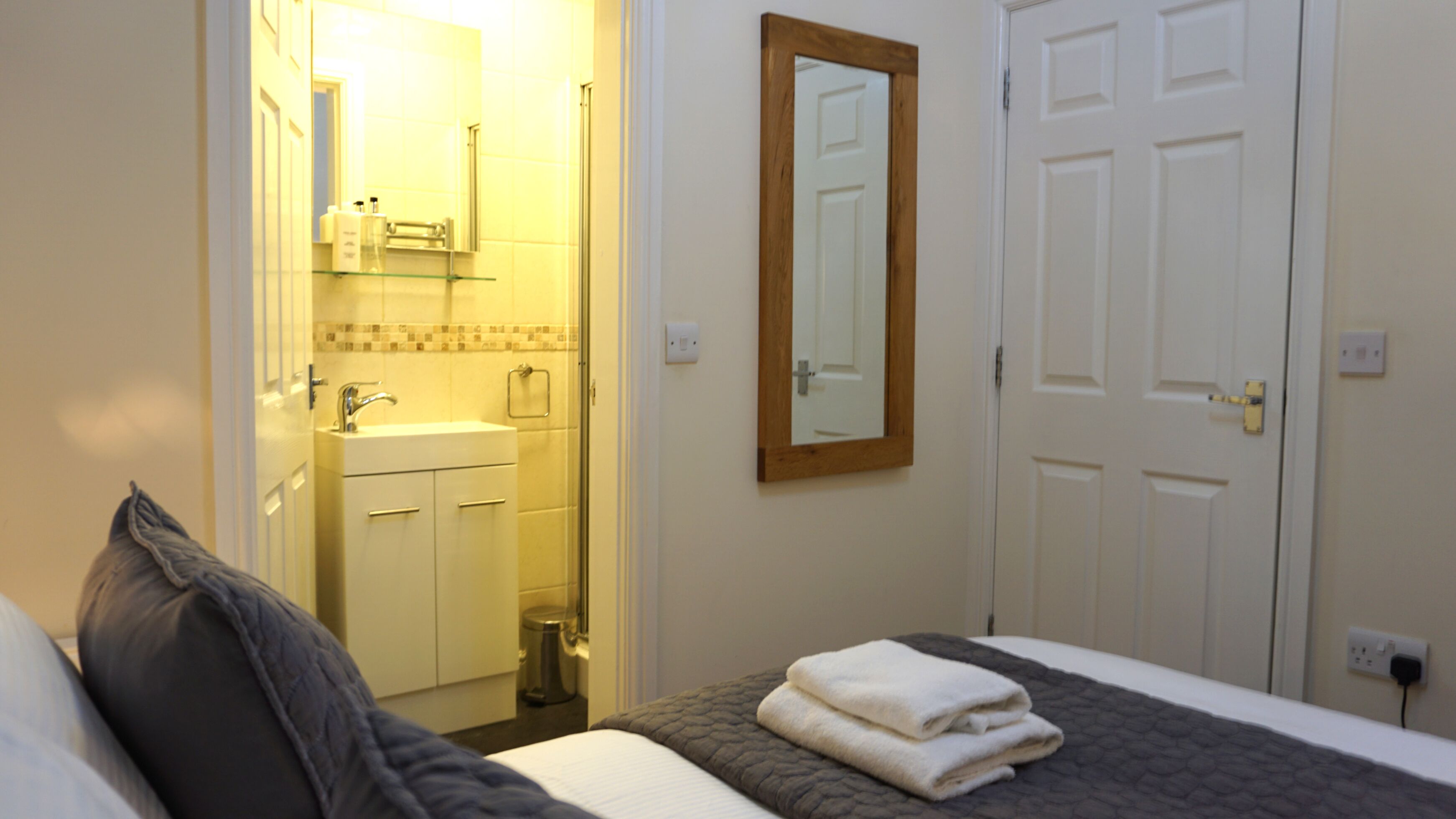 Ein Schlafzimmer mit Bett, Nachttisch, Spiegel, einer Tür und einem Badezimmer mit Waschbecken und Handtuchhalter.