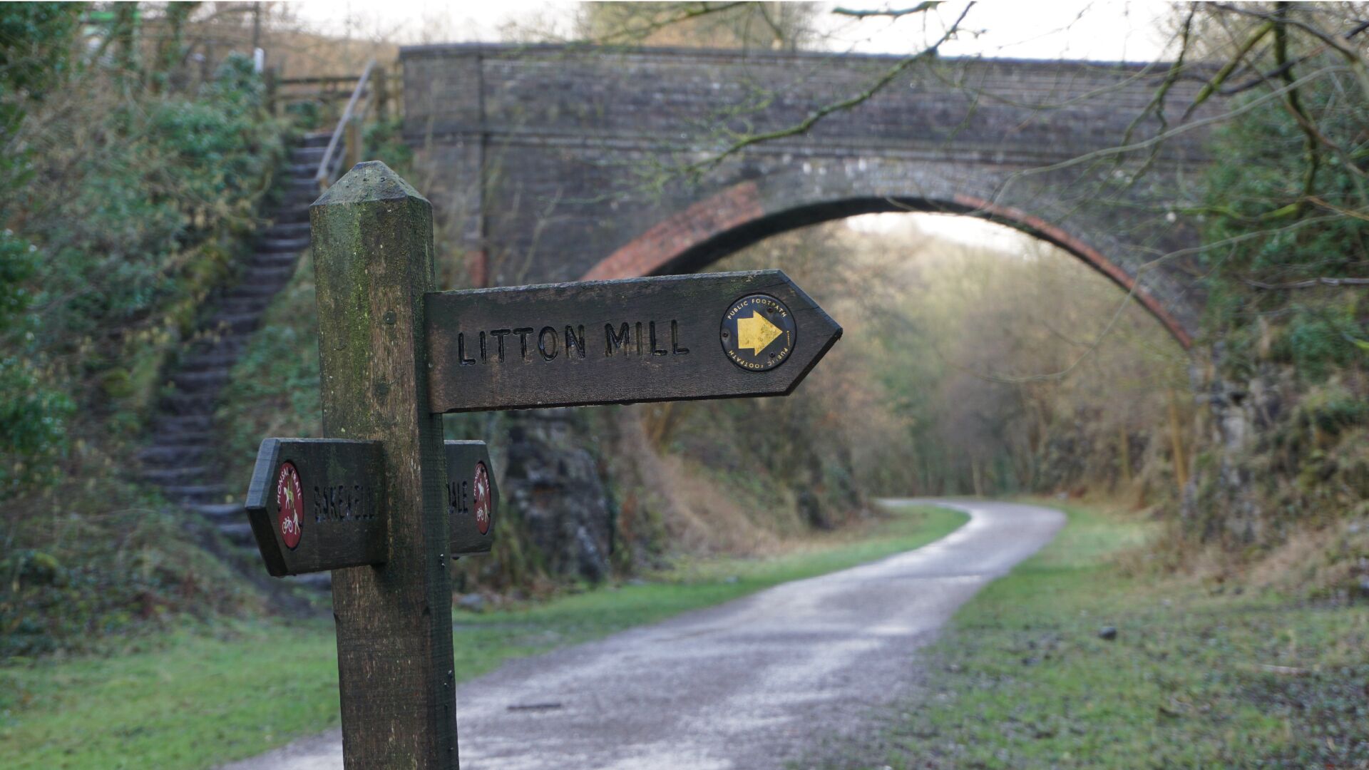 A directional signpost with 'LITTON MILL' pointing to the right, a brick bridge in the background, and a narrow road curving under the bridge.