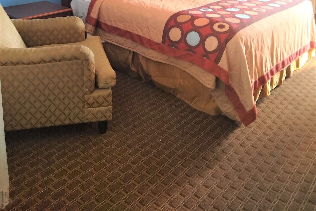Quarto de hotel com cama grande, cabeceira de madeira, poltrona estampada e janela com cortinas.