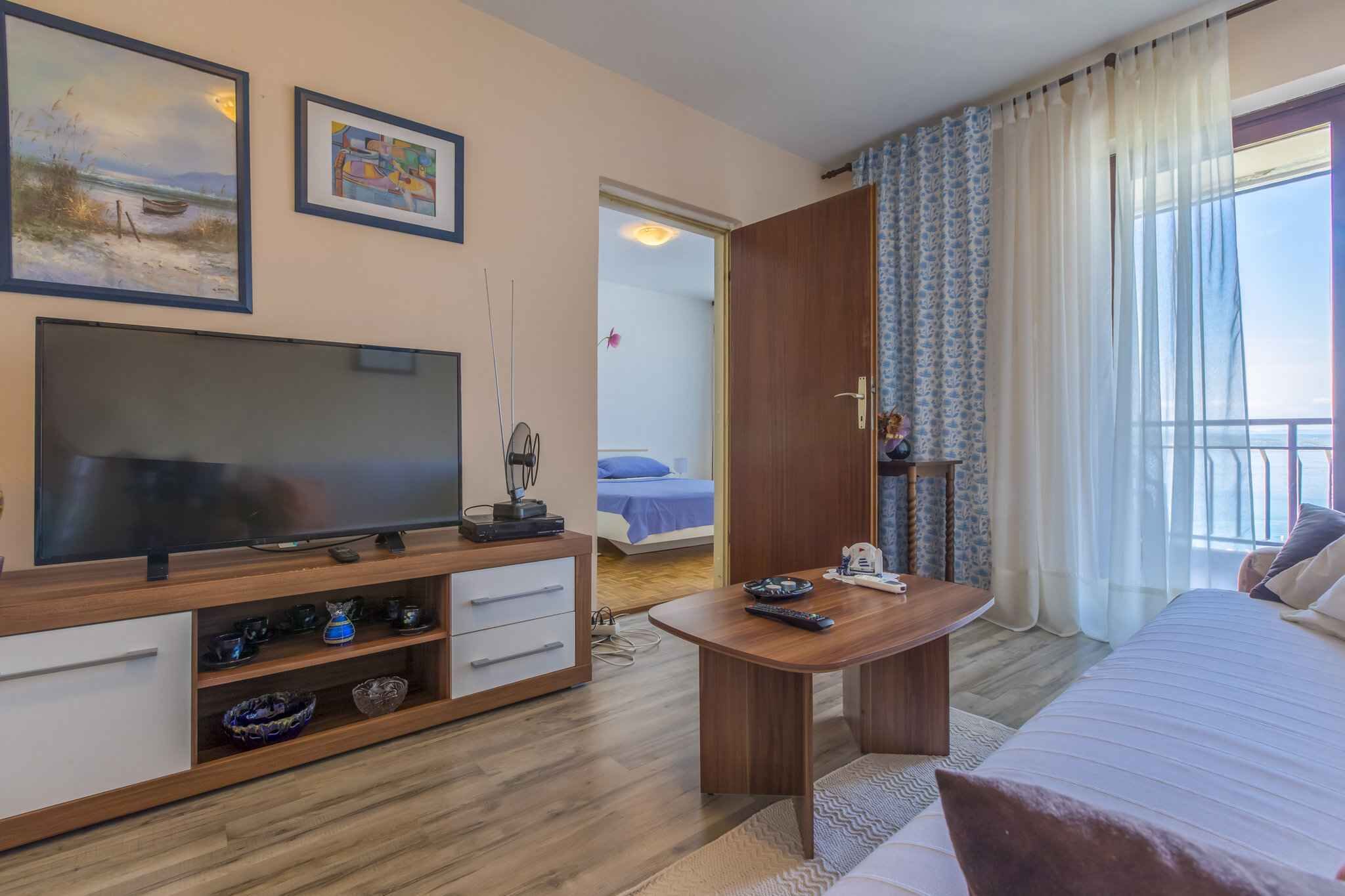 A hotel room with a TV, a wooden TV stand, a bed, a bedside table, a balcony with curtains, and artwork on the wall.