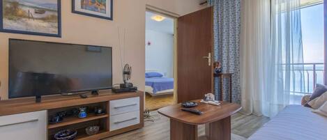 A hotel room with a TV, a wooden TV stand, a bed, a bedside table, a balcony with curtains, and artwork on the wall.