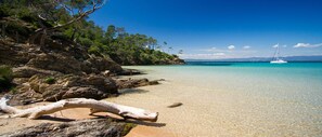 Une plage à l’eau turquoise et limpide, un voilier et un paysage rocheux parsemé d’arbres.