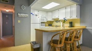 Cozinha compacta com armários brancos, micro-ondas, pia e um pequeno espaço bar com cadeiras de vime.