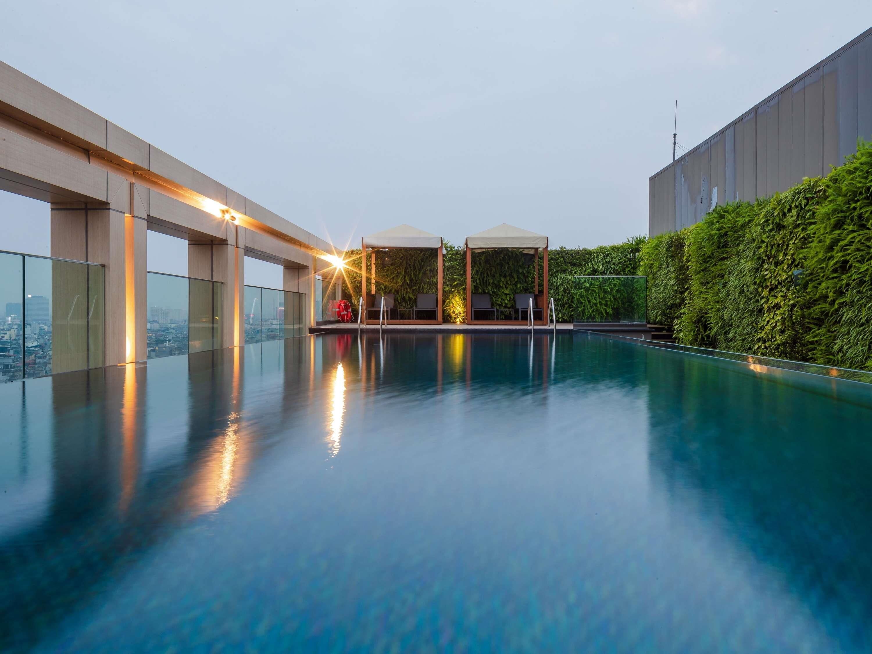 Una piscina infinita en la azotea con barandilla de cristal, un área de descanso cubierta con sillas y vistas a la ciudad.