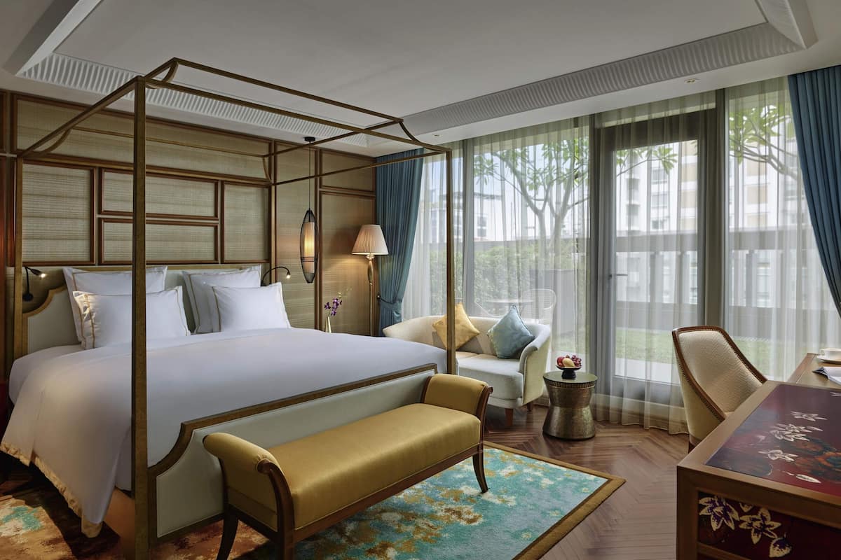 A spacious bedroom with a four-poster bed, a sitting area with a bench, a small table with a bowl of fruit, and a view of the outdoors through large windows.