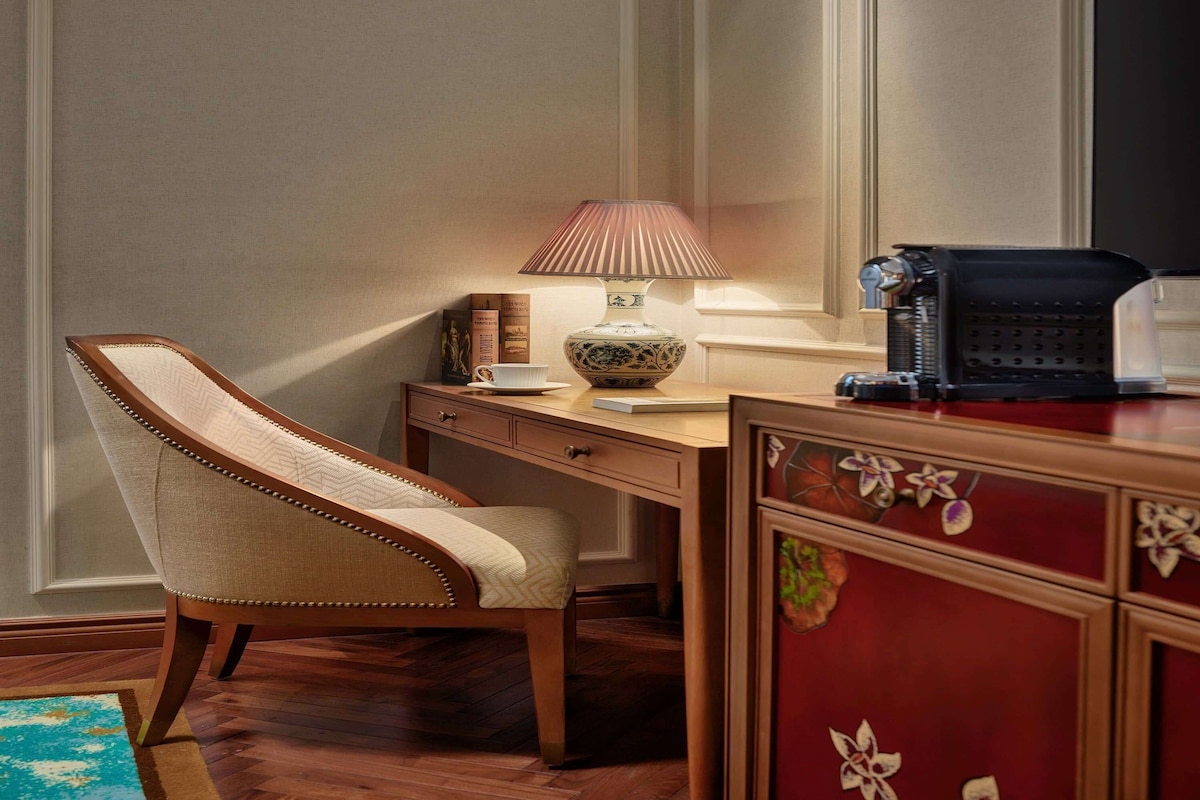A corner room with a wooden desk, a chair, a lamp, a coffee maker, and a decorative cabinet.