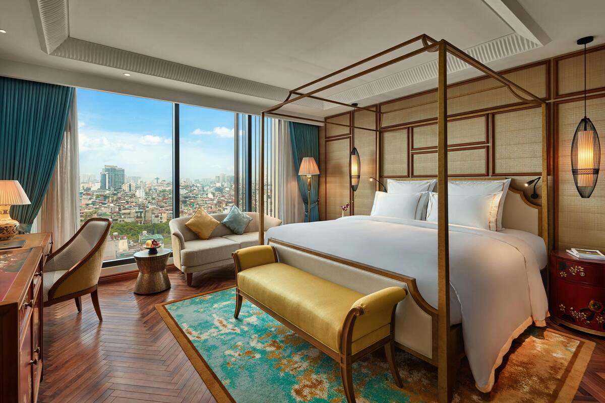 A spacious hotel room with a large bed, a sitting area with a sofa and armchair, a desk with a lamp, and a view of the cityscape through large windows.