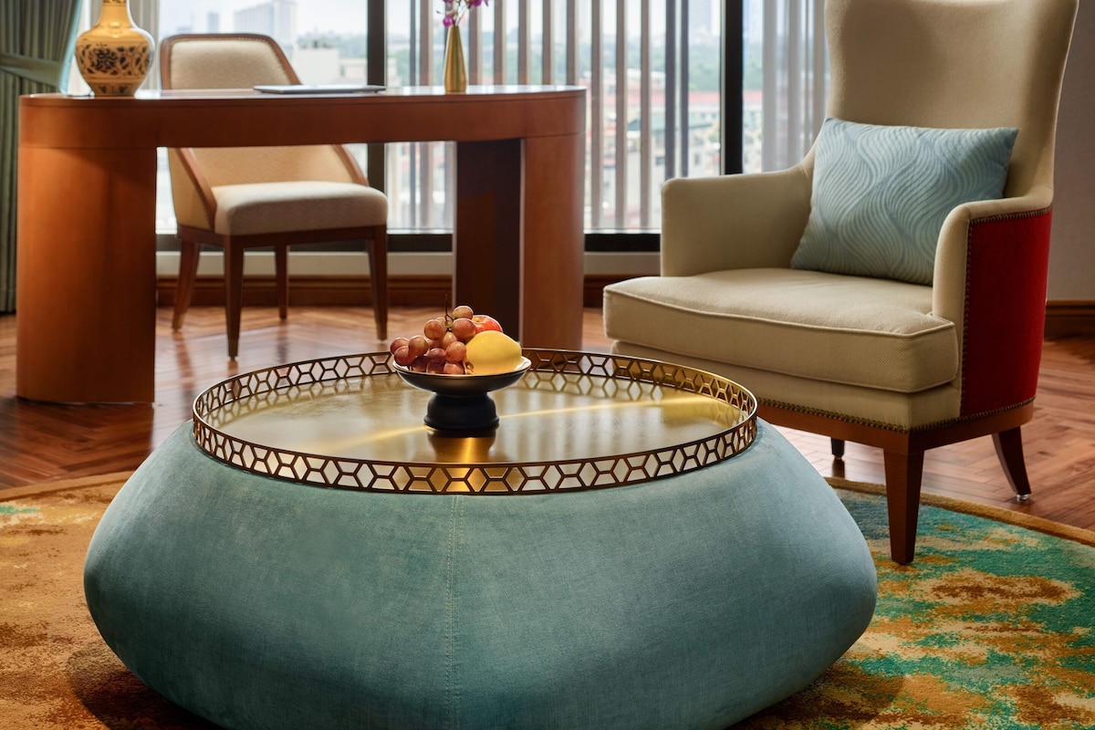 A modern hotel room with a large window, a round coffee table with fruits, a chair, and two lamps.