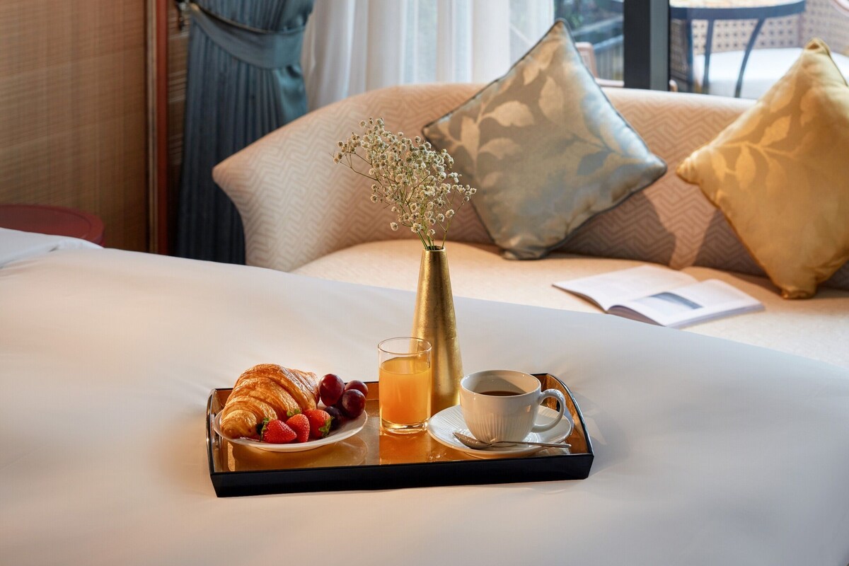 A neatly made bed with a tray of breakfast items, including croissants, strawberries, and orange juice, in a hotel room with a view of greenery outside.