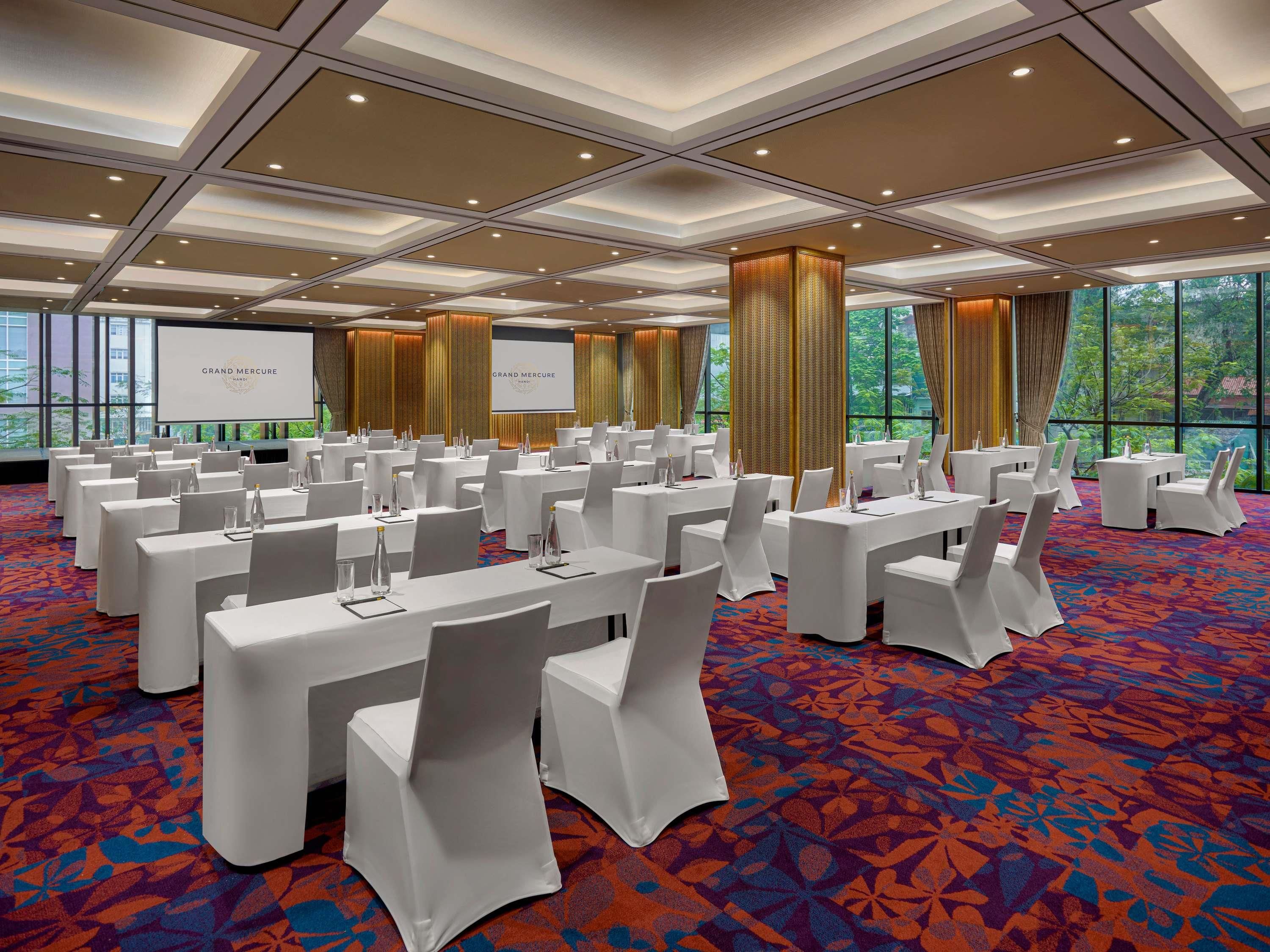 Un salón de conferencias con mesas, sillas y una pantalla grande que muestra 'GRAND MERCURE'.