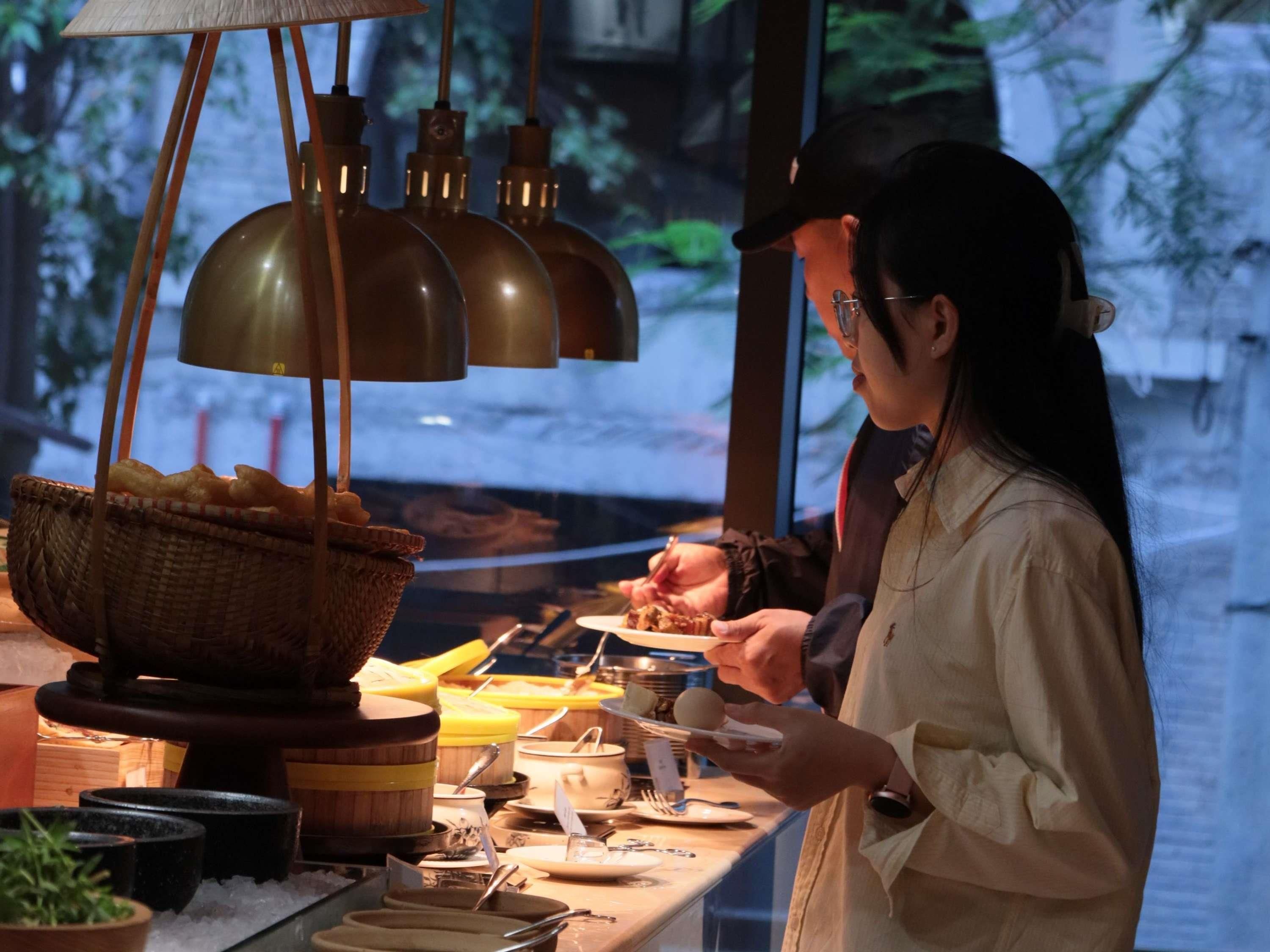 A person serving food at a counter with a basket of bread, a stove, and various kitchen utensils.