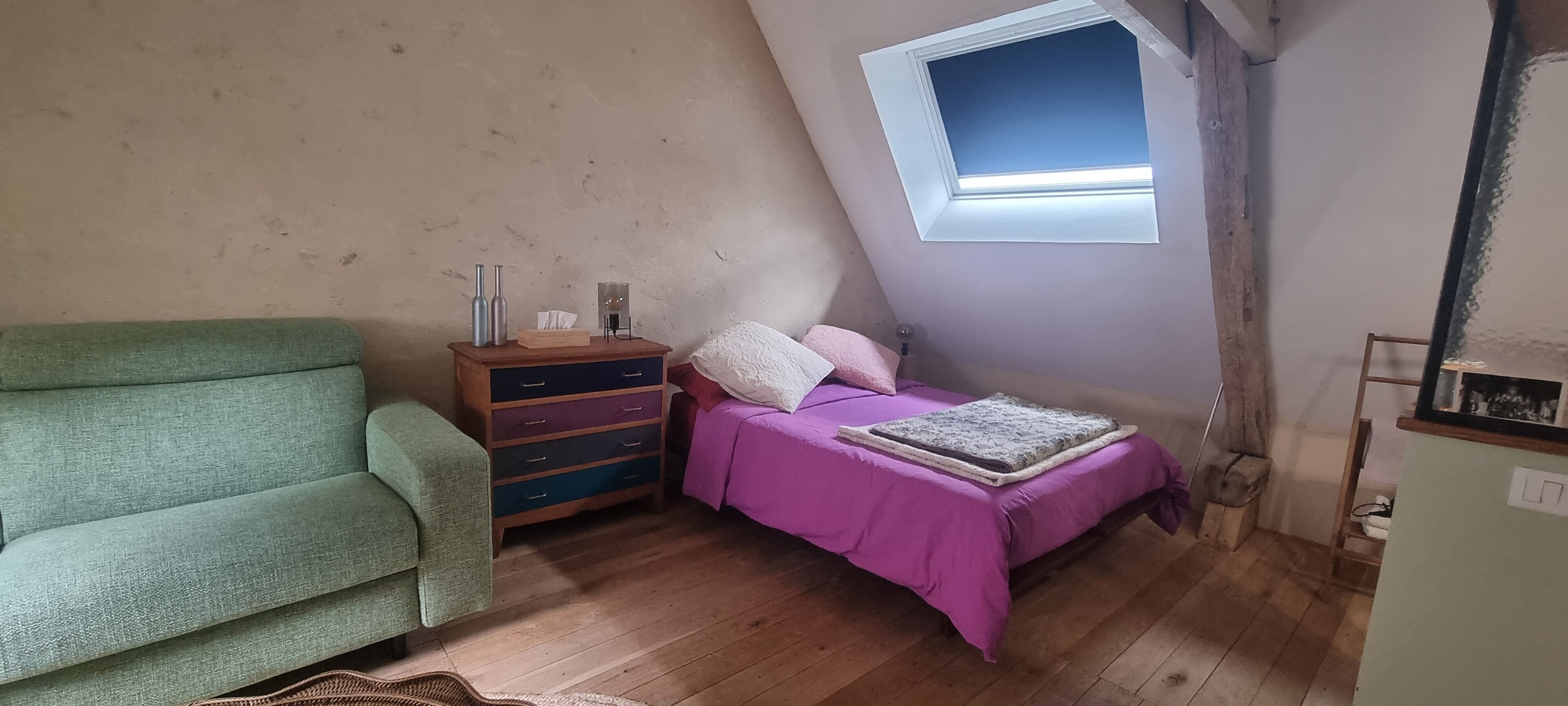 Une petite pièce agréable, avec un plafond en pente, une commode en bois dotée de tiroirs, un canapé vert, un lit recouvert d’une couverture violette et une lucarne.