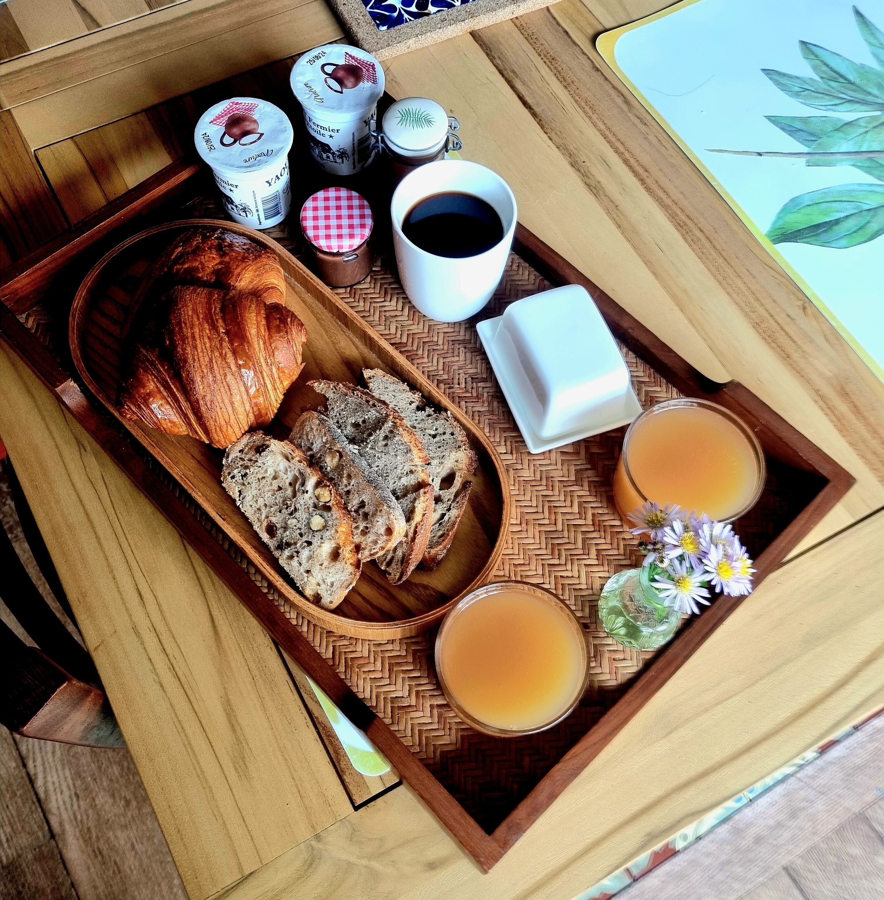 Ein Holztablett mit Croissants, Brot, Kaffee und Tee auf einer gewebten Matte.