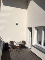Un balcón con una pequeña mesa, dos sillas, una ventana y una lámpara fijada a la pared.