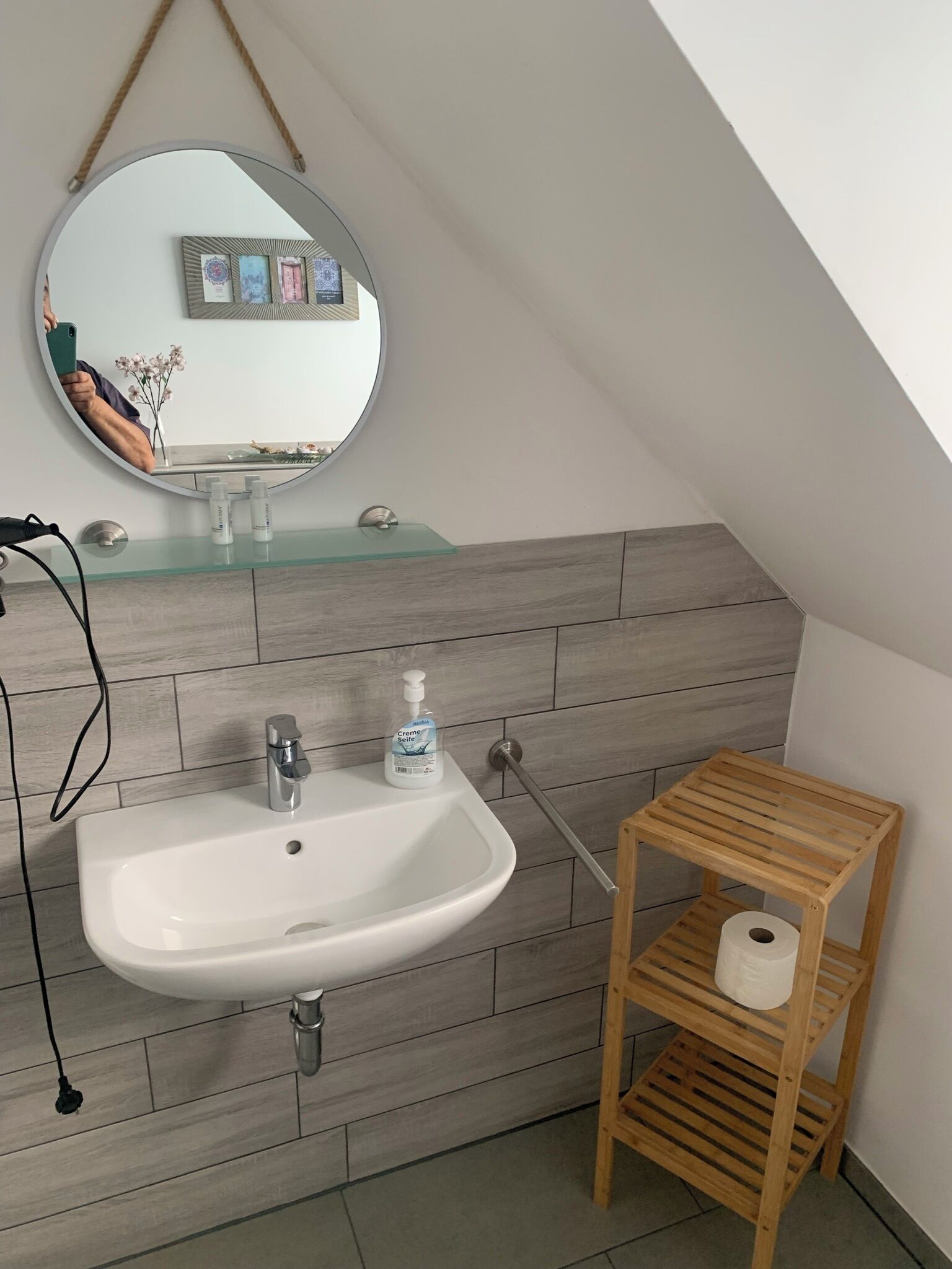 A bathroom with a white sink, a mirror, and a wooden shelf with a roll of toilet paper.
