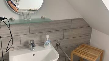 A bathroom with a white sink, a mirror, and a wooden shelf with a roll of toilet paper.
