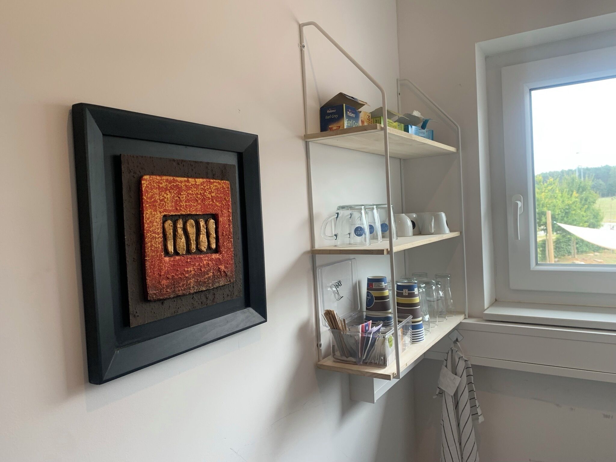 A room with a framed artwork on the wall, a shelf with various items, and a window with a view.