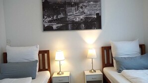 Habitación de hotel con dos camas, cada una con un cojín y una mesita de noche con una lámpara. Un cuadro de un paisaje urbano cuelga de la pared sobre las camas.