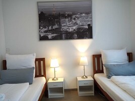 Habitación de hotel con dos camas, cada una con un cojín y una mesita de noche con una lámpara. Un cuadro de un paisaje urbano cuelga de la pared sobre las camas.