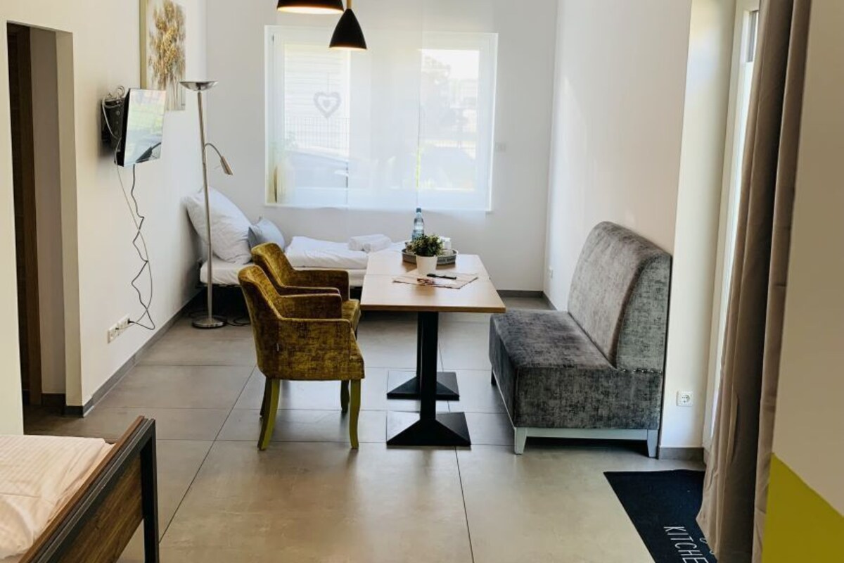 Una habitación moderna y bien iluminada con una cama, un sofá, una mesa de comedor y una silla. Hay una ventana con vistas al exterior.