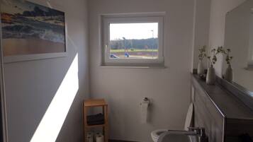 A bathroom with a window, a wooden shelf with towels, a toilet, and a mirror.