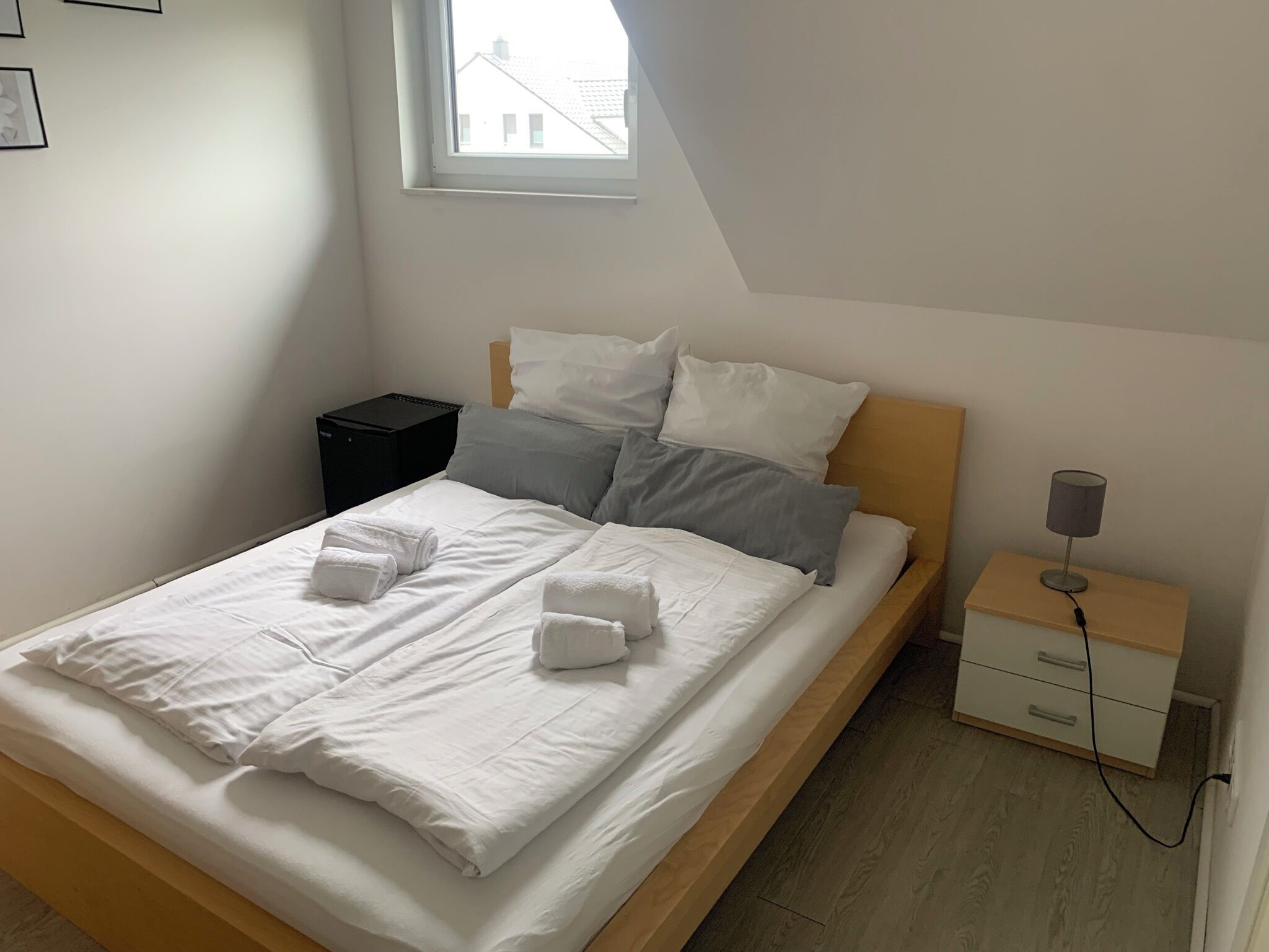 A neatly made bed with white linens and two rolled towels, a bedside table with a lamp, and a small nightstand.