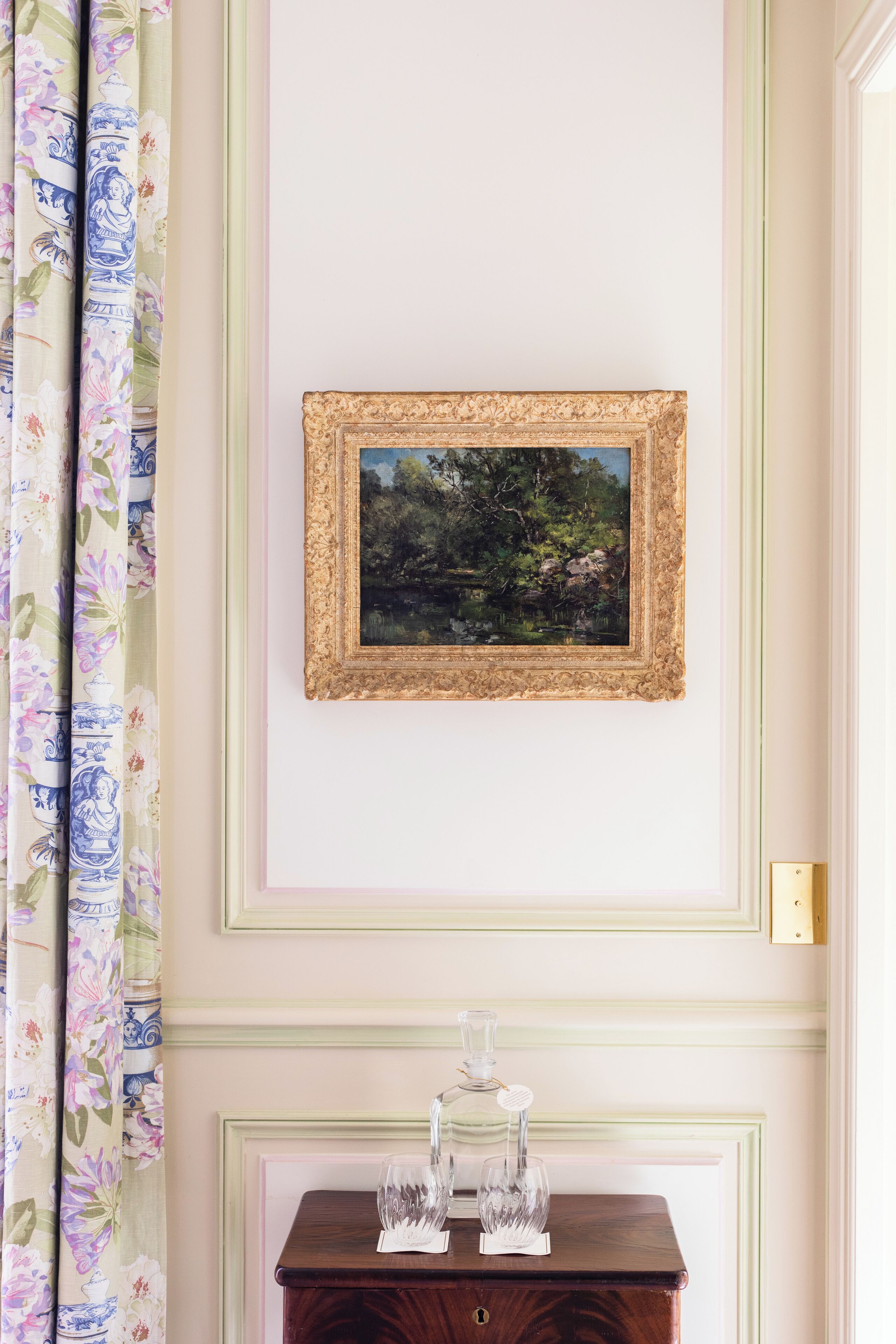 A framed landscape painting on a wall, a wooden side table with glassware, and floral curtains.