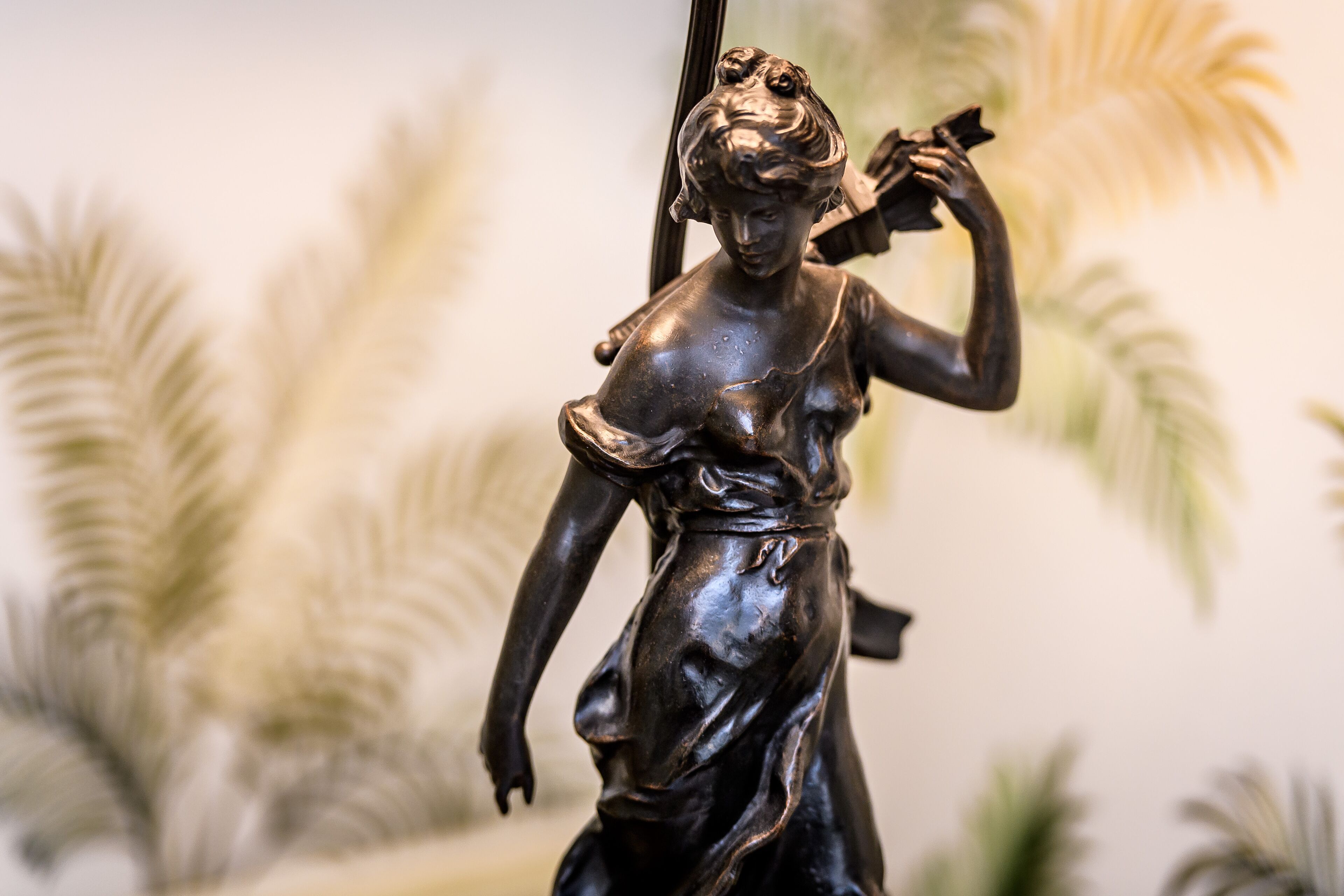 En bronze statue af en kvinde, der holder en blomst, foran en sløret baggrund med palmeblade.