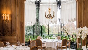 Un comedor con mesas dispuestas para una comida, rodeado de paneles de madera y grandes ventanales con cortinas.