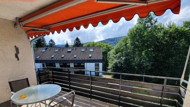 Un balcon avec une table et des chaises en verre, un auvent rouge et une vue sur un immeuble équipé de panneaux solaires.