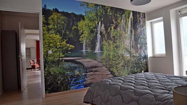 Ein Schlafzimmer mit einer großen Wandmalerei eines Waldwasserfalls, einem Bett mit grauer Steppdecke und Holzboden.
