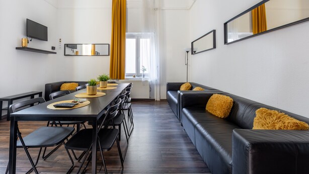 Een moderne woonkamer met een zwarte leren bank, een eettafel met stoelen, een flatscreen televisie en een groot raam met gordijnen.