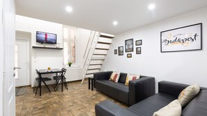 Een moderne woonkamer met een zwarte leren bank, een eettafel met stoelen, een aan de muur bevestigde televisie, en een trap die naar een ander niveau leidt.