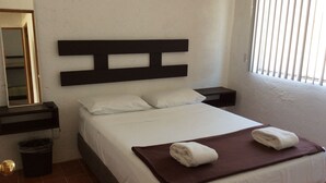 Una cama bien hecha con sábanas blancas, una manta marrón y dos toallas dobladas. Hay una mesita de noche con una lámpara y un cesto de basura junto a la cama. Un espejo está montado en la pared sobre la cama.