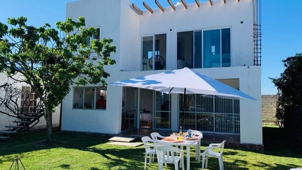 Una casa moderna de dos pisos con un patio que cuenta con una mesa, sillas, un paraguas blanco y un amplio ventanal con vista al jardín.